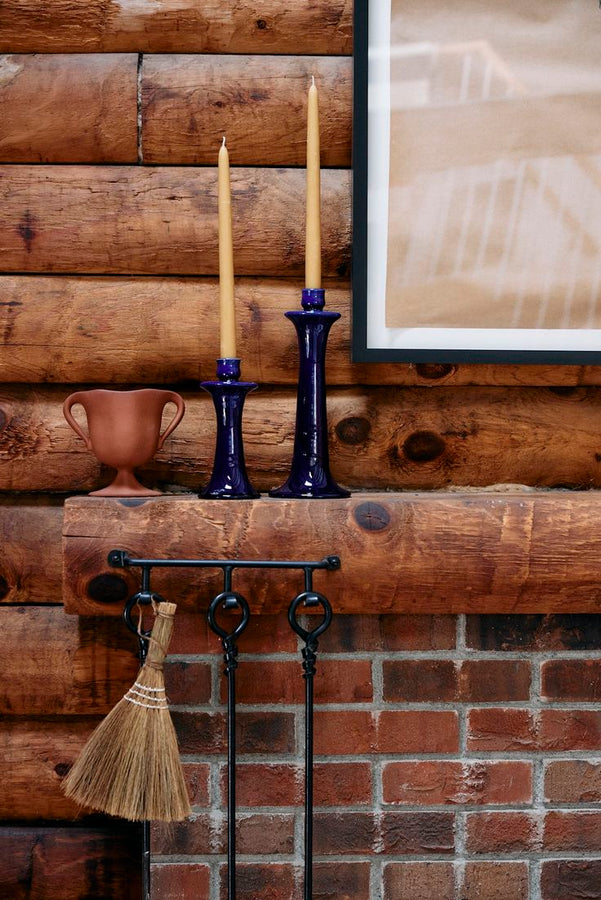 A wooden mantel displays two tall yellow candles in Maria Candleholders and a small brown vase, with fireplace tools and a broom hanging below against a brick and log wall.