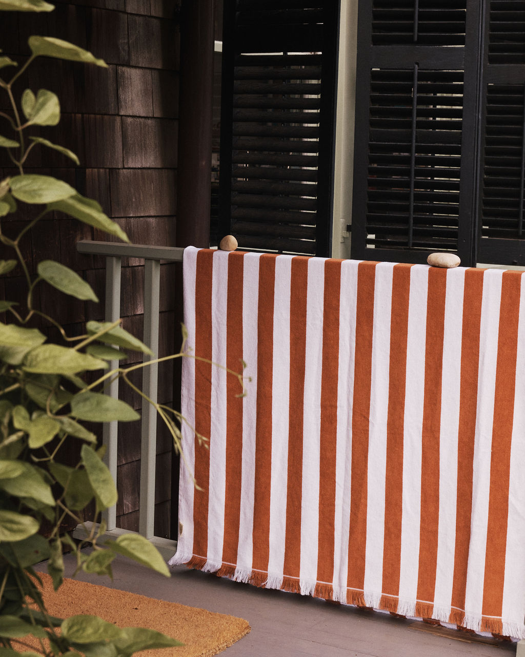 A Paloma Stripe Towel in red and white cotton drapes over a porch railing, secured with stones, beside a green plant and half-closed dark shutters.
