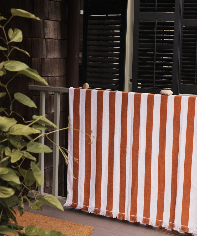 A Paloma Stripe Towel in red and white cotton drapes over a porch railing, secured with stones, beside a green plant and half-closed dark shutters.