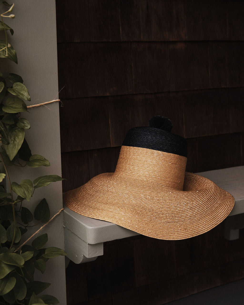 The Dahlia Hat, a handwoven straw garden hat with a black crown and elegant black ties, rests on a gray wooden bench beside brown shingled walls and lush green plants.