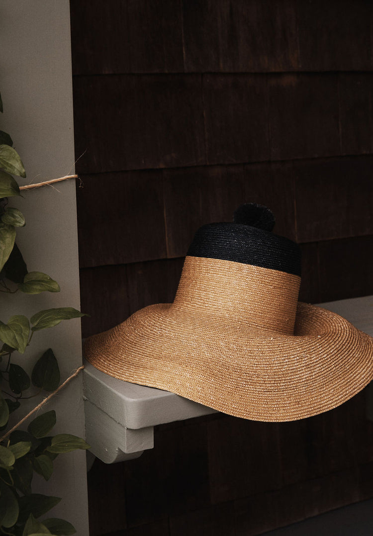 The Dahlia Hat, a handwoven straw garden hat with a black crown and elegant black ties, rests on a gray wooden bench beside brown shingled walls and lush green plants.