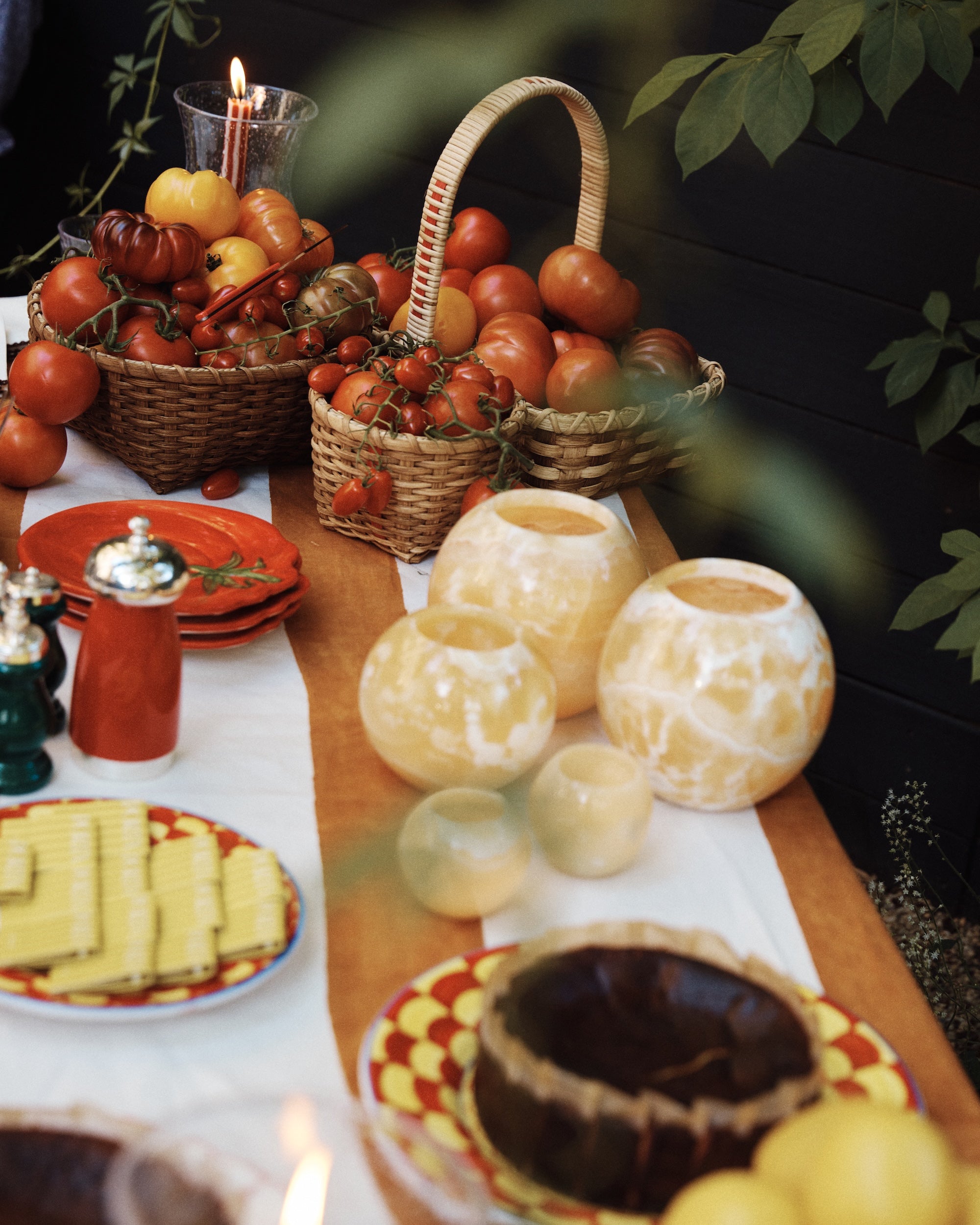 A table set outdoors with green leaves, featuring baskets of tomatoes, plates stacked with cheese slices, a round cake with a lit candle, and the unique Akila Candleholder.
