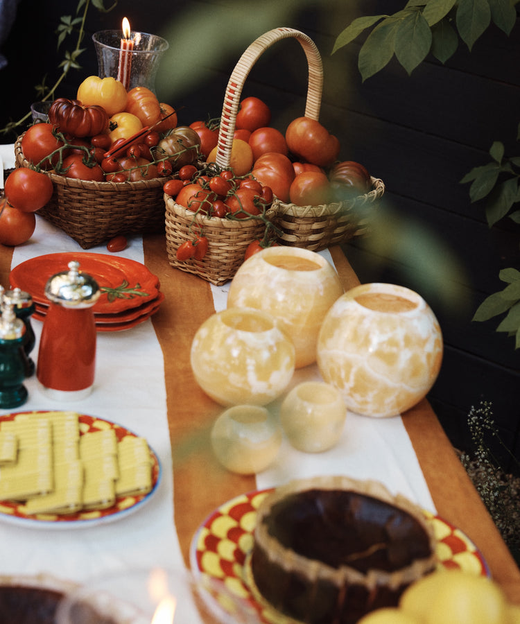 A table set outdoors with green leaves, featuring baskets of tomatoes, plates stacked with cheese slices, a round cake with a lit candle, and the unique Akila Candleholder.