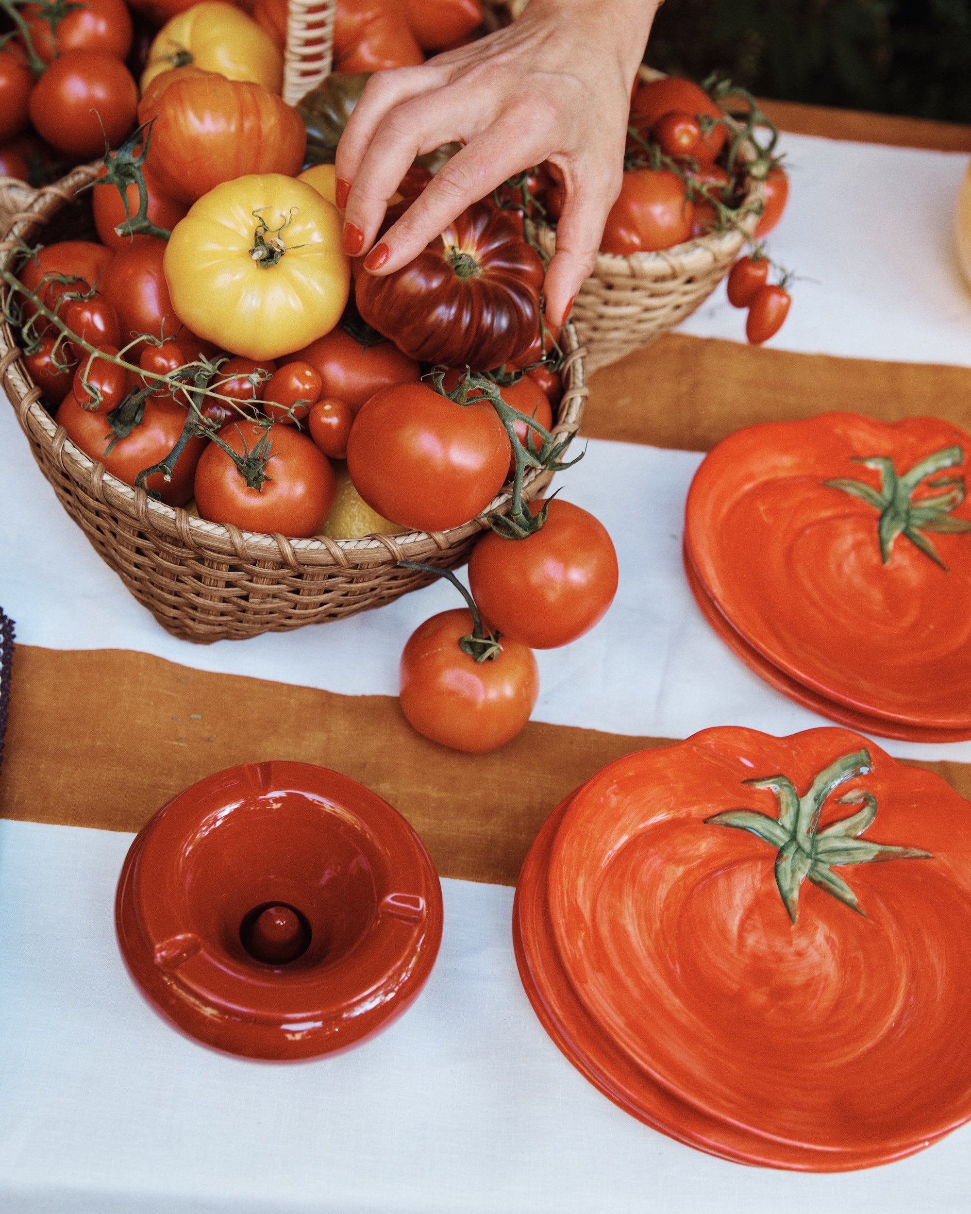 A hand reaches for a tomato in a basket of assorted tomatoes beside the Tomato Small Plate and a small dish on a striped tablecloth, highlighting vibrant Mediterranean tableware.