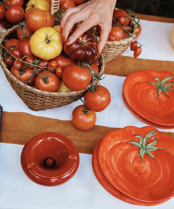 A hand reaches for a tomato in a basket of assorted tomatoes beside the Tomato Small Plate and a small dish on a striped tablecloth, highlighting vibrant Mediterranean tableware.