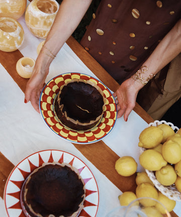 Someone in a brown dress places a dark round cake on the Palio Dinner Plate, The Snail, beside another cake, lemons, and ceramics on a table.