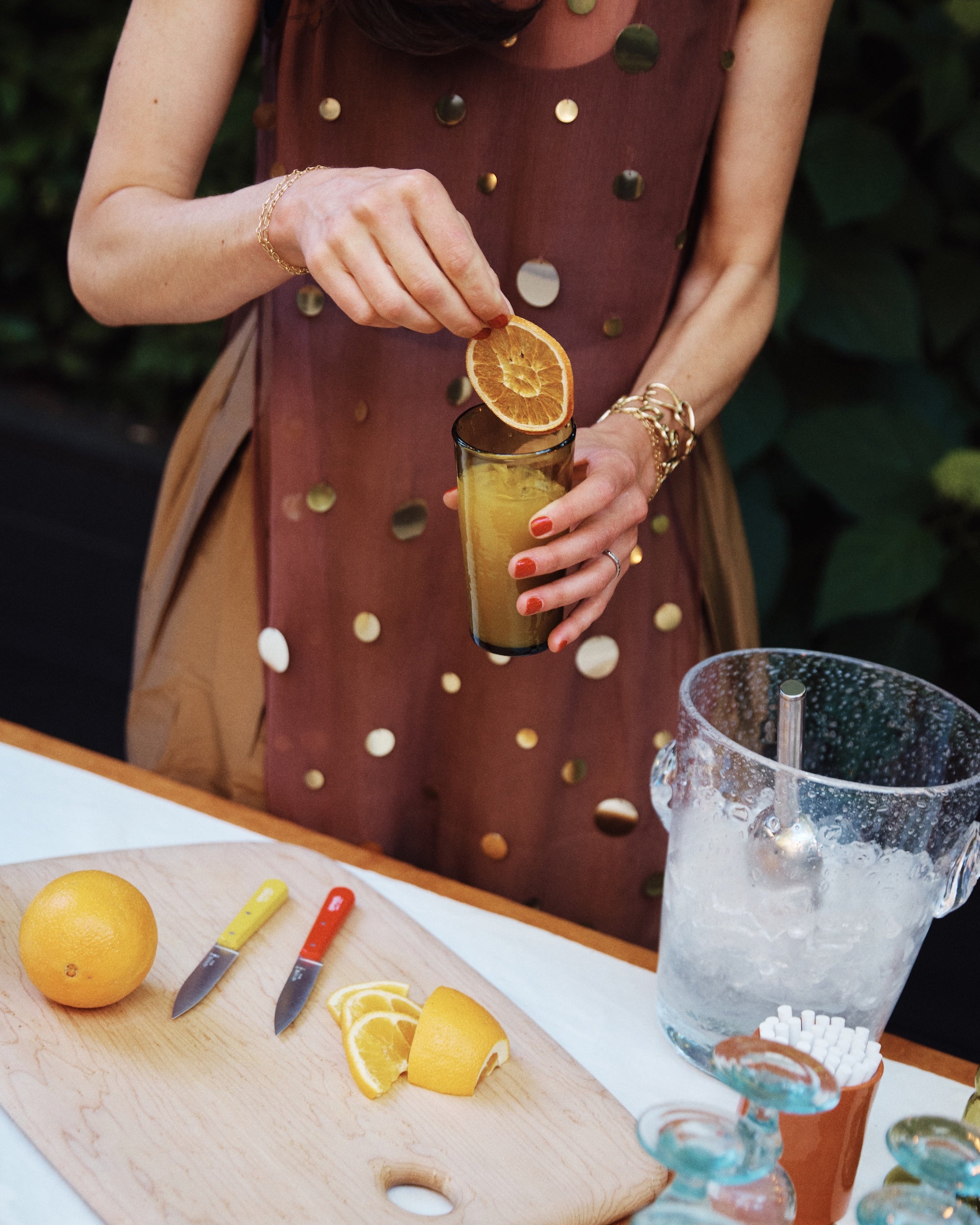A person garnishes an orange drink in elegant French glassware, placing it beside a cutting board with sliced oranges, two knives, and the Lauren Champagne Bucket.