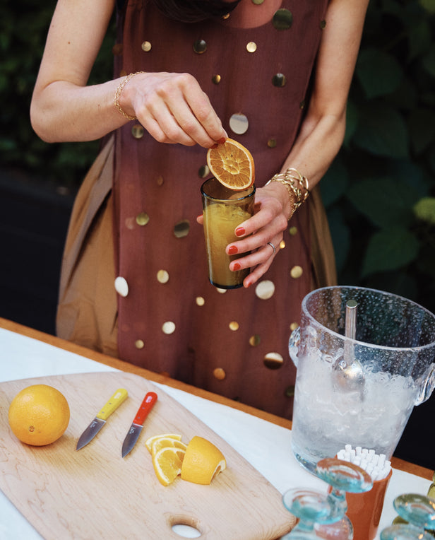 A person garnishes an orange drink in elegant French glassware, placing it beside a cutting board with sliced oranges, two knives, and the Lauren Champagne Bucket.