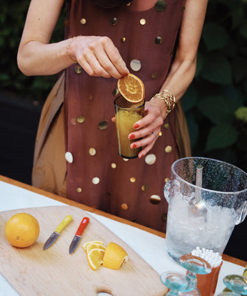 A person garnishes an orange drink in elegant French glassware, placing it beside a cutting board with sliced oranges, two knives, and the Lauren Champagne Bucket.