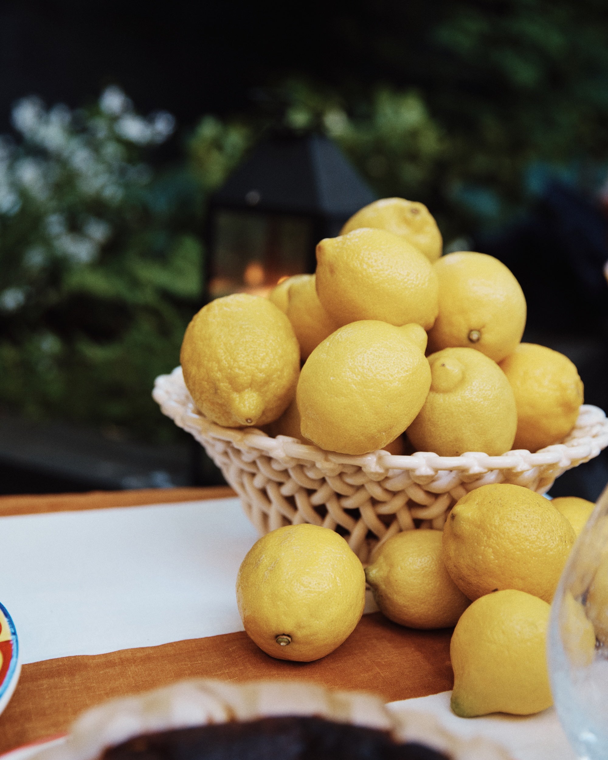 The Remi Decorative Fruit Bowl, filled with yellow lemons and placed on a table among more lemons, stands out as greenery and a lantern softly blur in the background, adding sculptural charm to the scene.