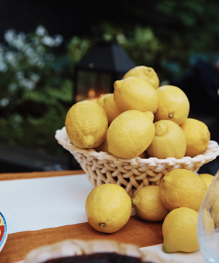 The Remi Decorative Fruit Bowl, filled with yellow lemons and placed on a table among more lemons, stands out as greenery and a lantern softly blur in the background, adding sculptural charm to the scene.