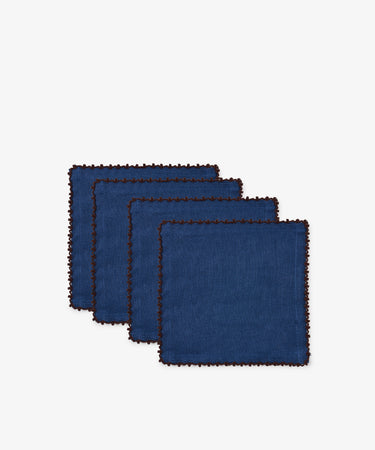 Set of four Nico Cocktail Napkins in indigo blue linen, each hand-finished with brown stitched edges, displayed in a staggered row on a white background.