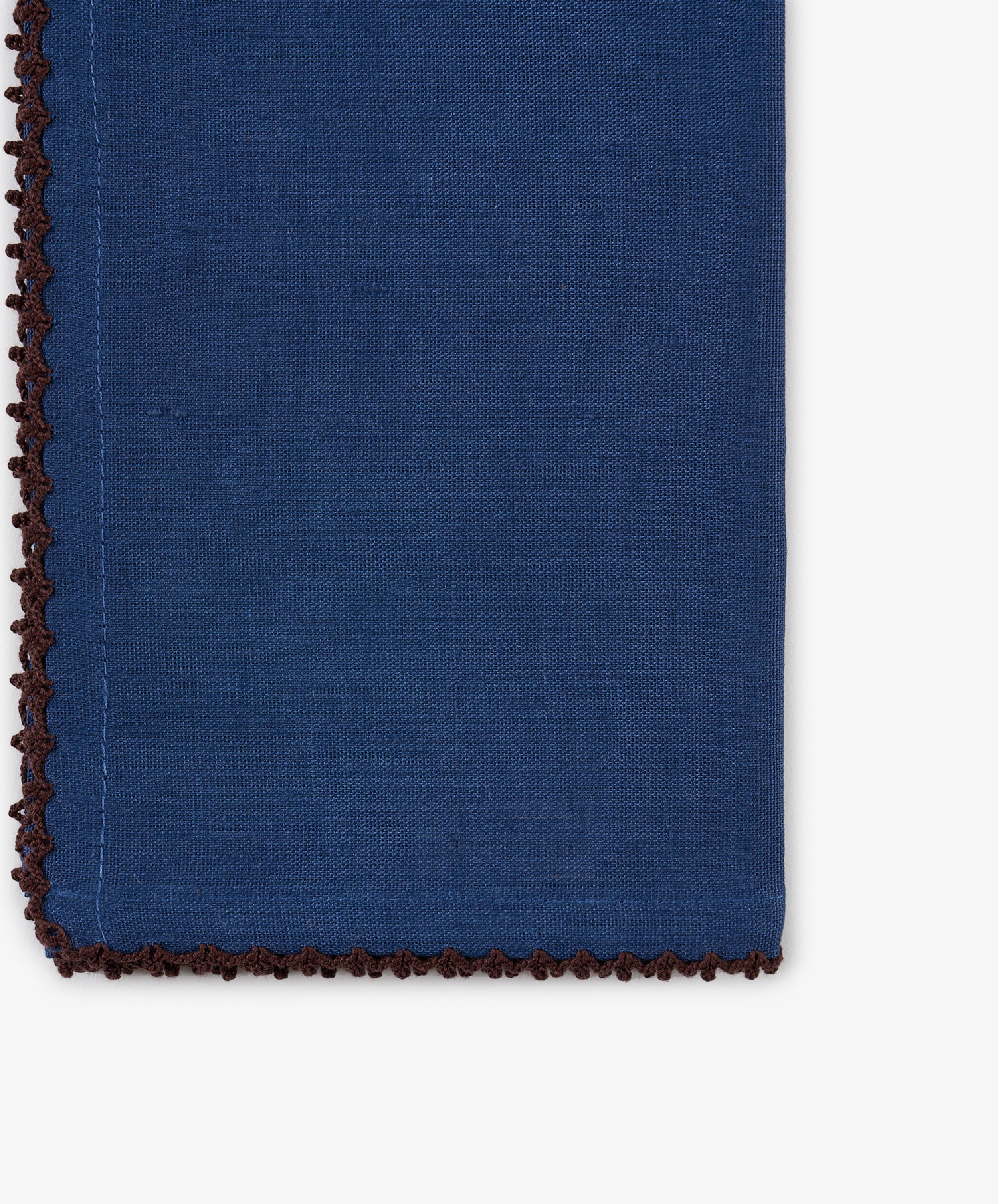 Close-up of the Nico Napkin in indigo blue linen, featuring a brown decorative stitched edge and one finished corner set against a white background. This hand-finished napkin from the set of 6 showcases classic craftsmanship and elegant detailing.