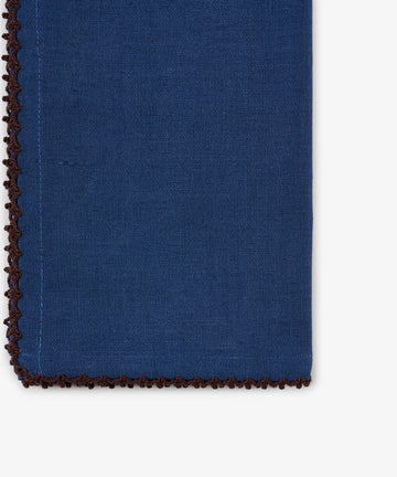 Close-up of the Nico Napkin in indigo blue linen, featuring a brown decorative stitched edge and one finished corner set against a white background. This hand-finished napkin from the set of 6 showcases classic craftsmanship and elegant detailing.