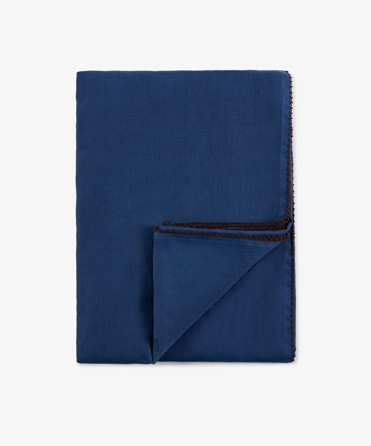 A neatly folded Nico Tablecloth in indigo blue with hand-finished brown stitching along the edges, displayed on a plain white background.