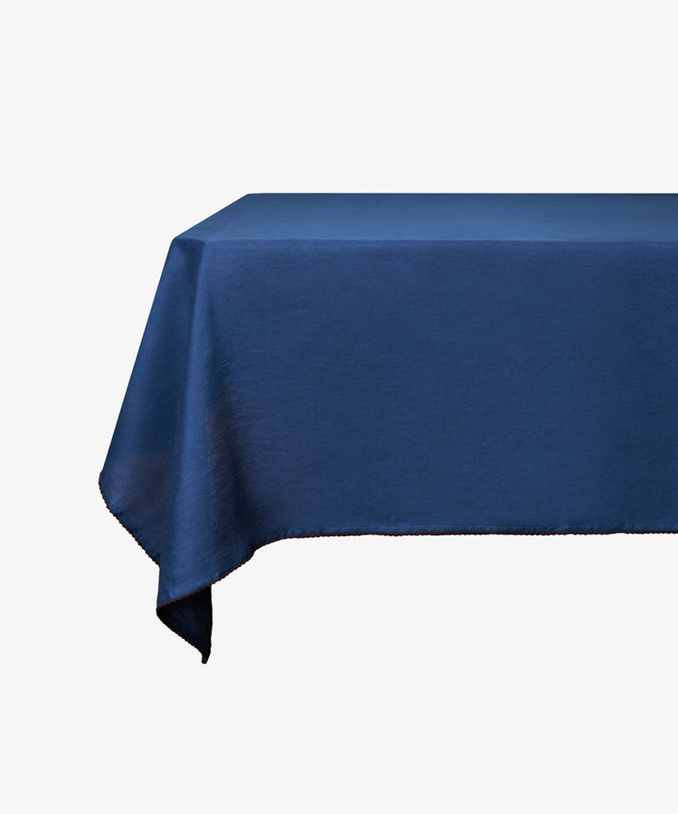 A rectangular table covered with the Nico Tablecloth, a hand-finished linen piece in indigo blue, set against a white background.