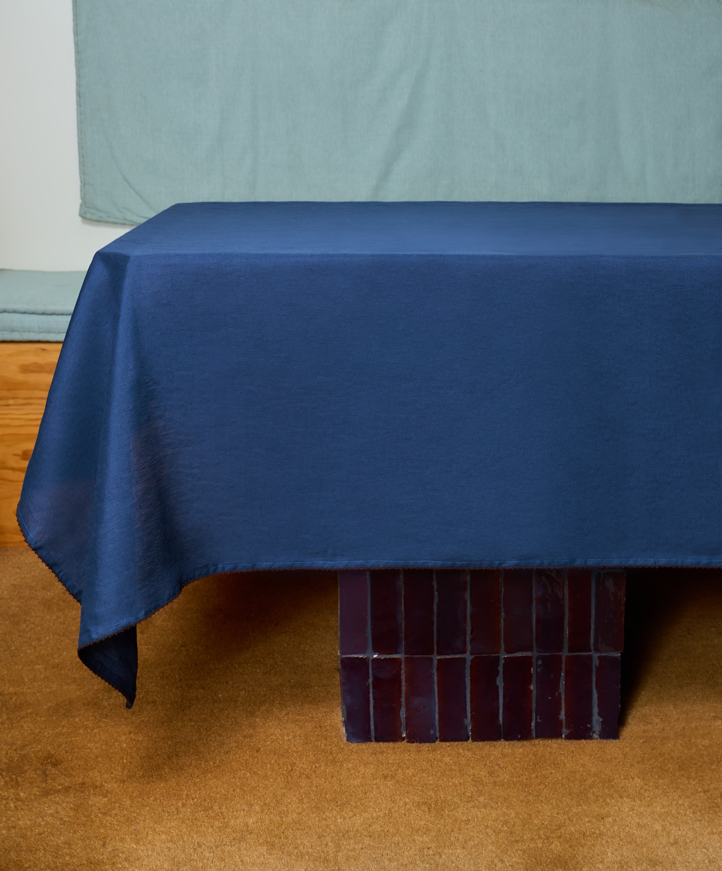 A rectangular table draped with the hand-finished Nico Tablecloth in indigo blue linen stands on a brown carpet, with a blue fabric background behind it.