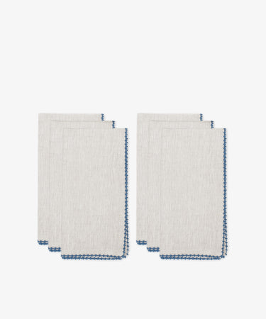 Six folded Nico Napkins in beige linen with blue stitched edges are stacked against a white background, perfect for elegant dining or as stylish modern table linens.