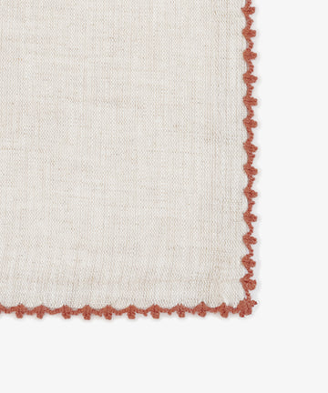 A close-up of the Nico Napkin, Set of 6, shows its beige linen fabric with a textured surface, decorative scalloped red border, and contrasting edge stitching.