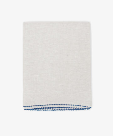 The Nico Tablecloth, in light beige linen with blue stitching along one edge, is neatly folded and hand-stitched, shown against a plain white background.