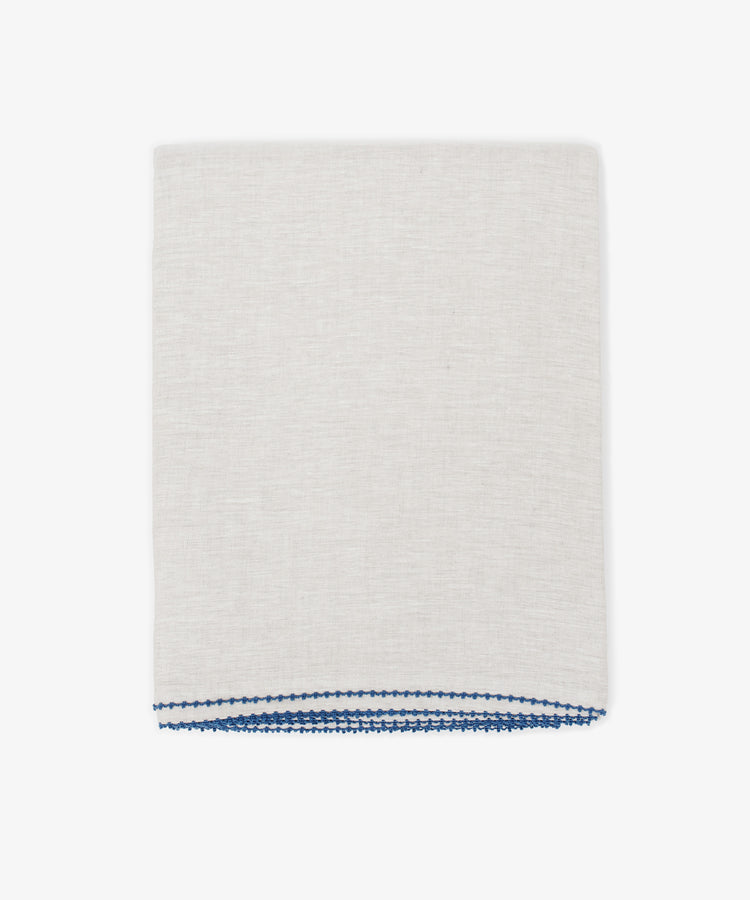 The Nico Tablecloth, in light beige linen with blue stitching along one edge, is neatly folded and hand-stitched, shown against a plain white background.