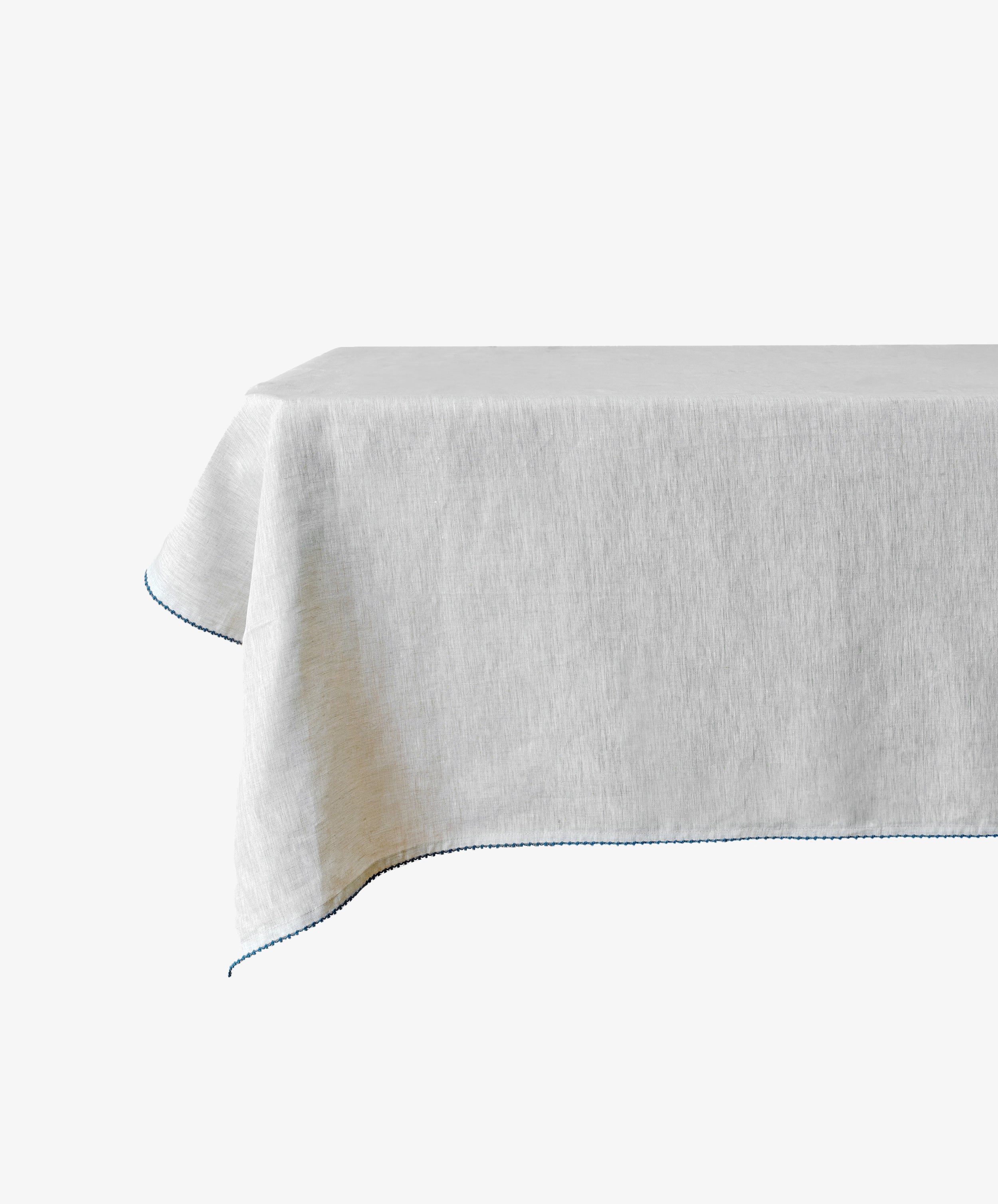 The Nico Tablecloth is a rectangular, plain white linen tablecloth with a hand-stitched blue edge—ideal for elegant table decor—shown on a table against a white background.