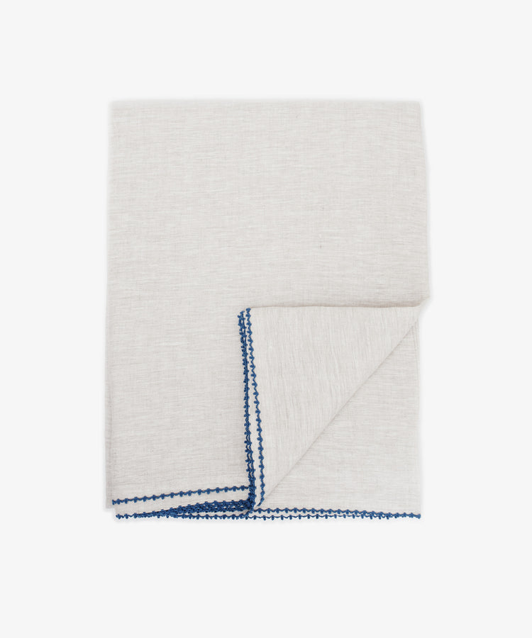 The Nico Tablecloth, in light gray with blue hand-stitched edging for elegance, is displayed folded on a white background.