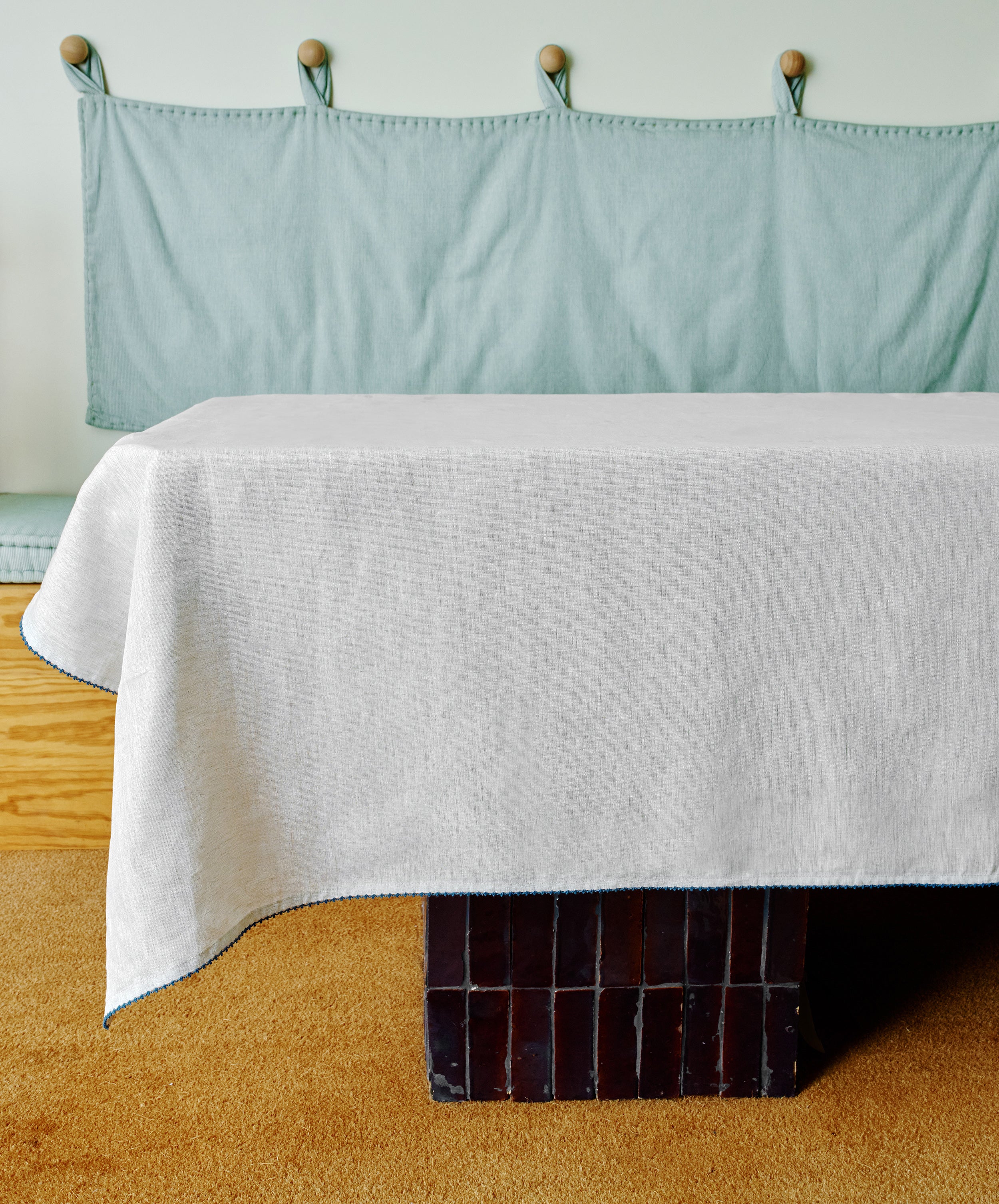 The Nico Tablecloth, hand-stitched from white linen, covers a rectangular table in a room with a light blue cushioned bench, wooden accents, and a beige carpet.