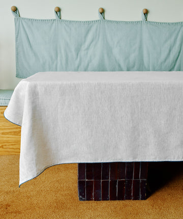 The Nico Tablecloth, hand-stitched from white linen, covers a rectangular table in a room with a light blue cushioned bench, wooden accents, and a beige carpet.