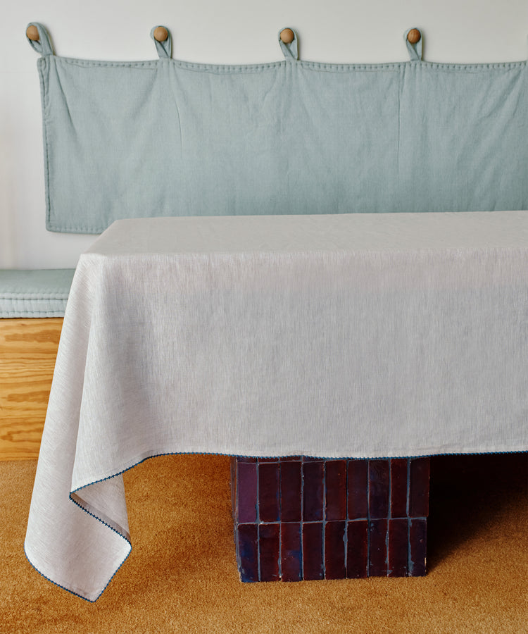 A Nico Tablecloth in light linen covers a table on a brown tile base, with a padded bench and light blue wall cushion behind it.