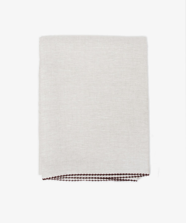The Nico Tablecloth features light-colored, subtly textured fabric with dark hand-stitched detailing along the edge—perfect for elegant table linens.