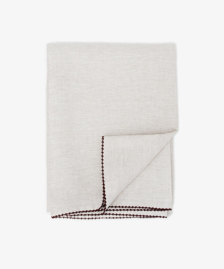 The Nico Tablecloth is a neatly folded light beige linen tablecloth with dark brown hand-stitched edges, displayed on a white background.