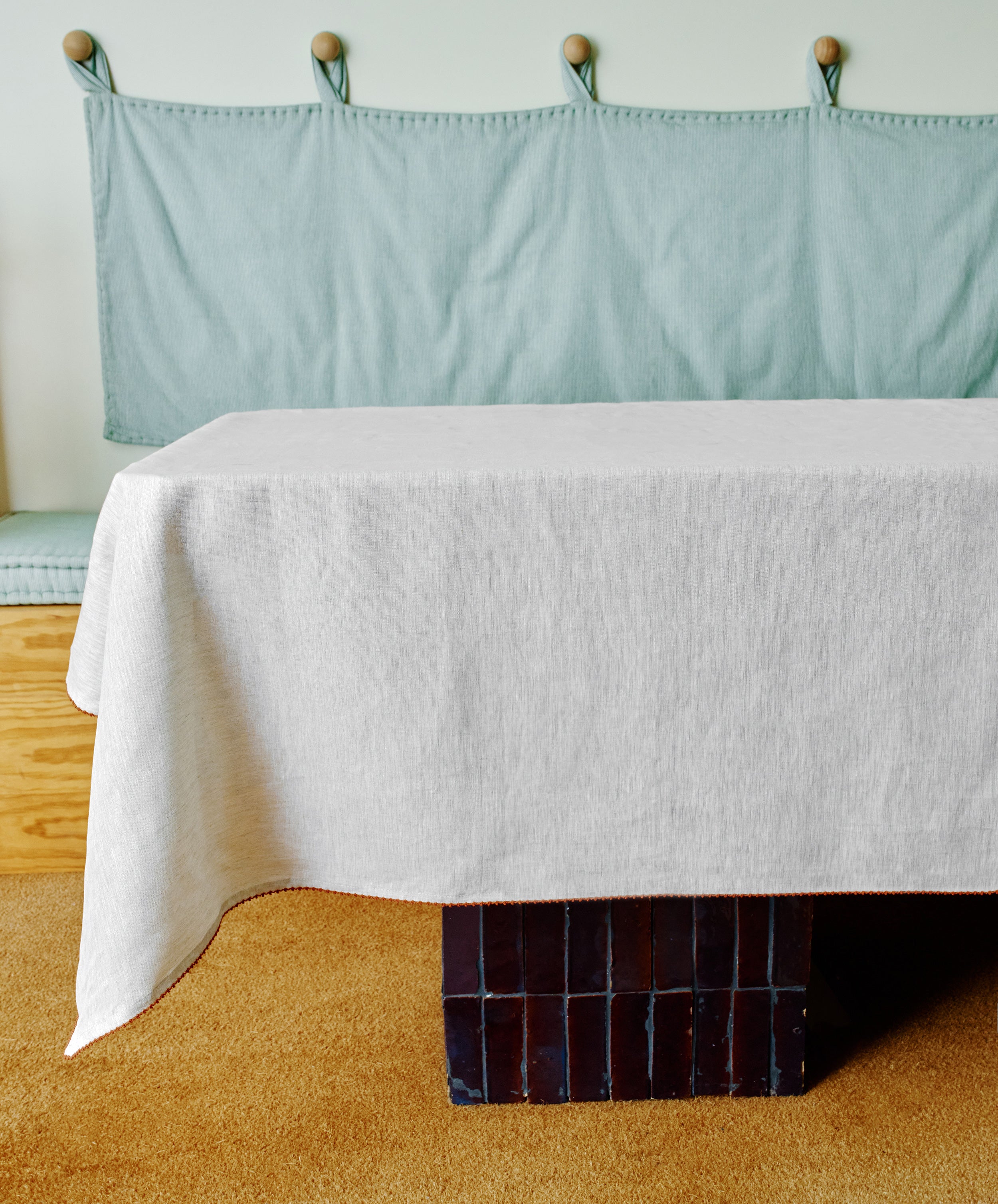The Nico Tablecloth, hand-stitched from white linen, drapes over a rectangular table with a black tiled base, set in front of a light blue cushioned bench on a tan carpet.