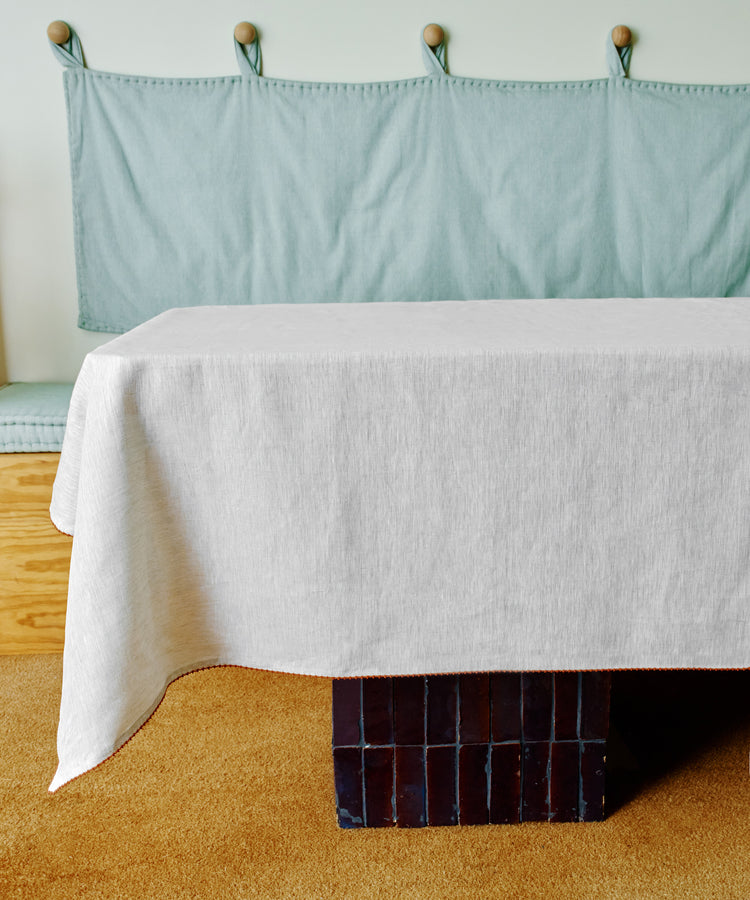 The Nico Tablecloth, hand-stitched from white linen, drapes over a rectangular table with a black tiled base, set in front of a light blue cushioned bench on a tan carpet.