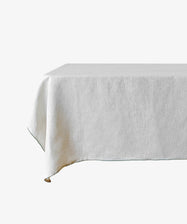 The Nico Tablecloth, a plain white linen piece, is draped over a rectangular table against a white background, bringing a touch of elegance to classic table linens.
