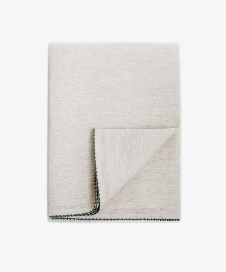 The Nico Tablecloth is shown folded in light beige, featuring visible hand-stitched edging and a flipped corner that reveals the underside.