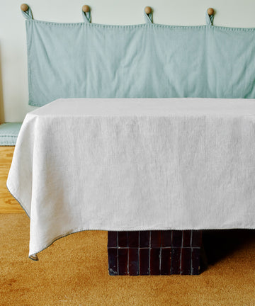 A rectangular table draped with the Nico Tablecloth sits before a cushioned bench with a light blue cover, on a tan carpeted floor.