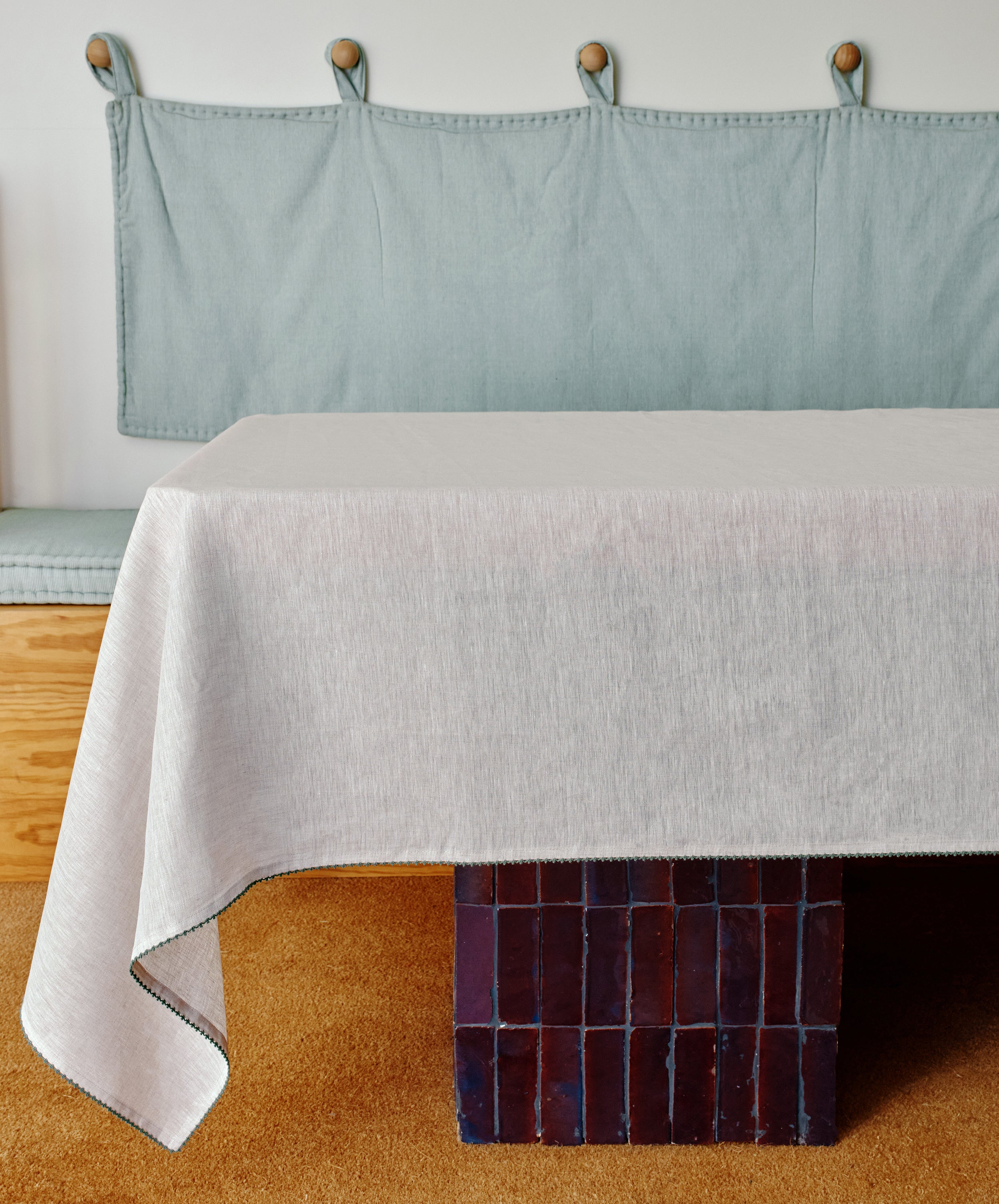 The Nico Tablecloth in light grey linen with hand-stitched detail covers a rectangular table with a dark tiled base. In the background, there’s a cushioned bench featuring a fabric backrest and wood accents.