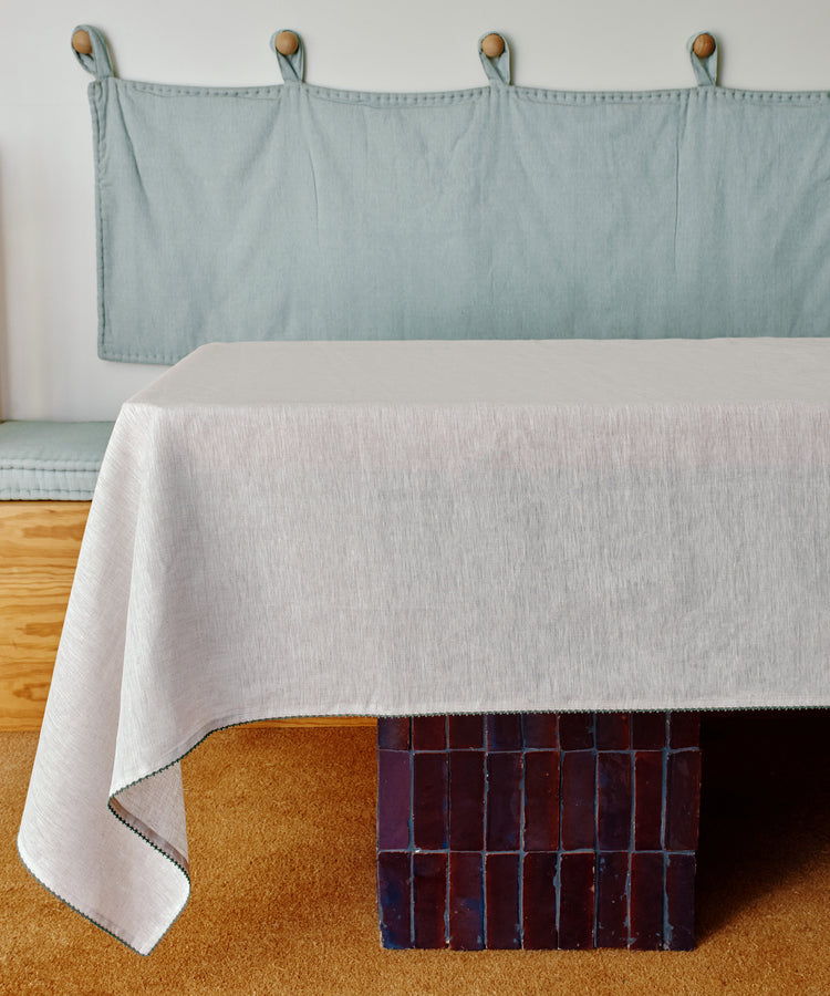 The Nico Tablecloth in light grey linen with hand-stitched detail covers a rectangular table with a dark tiled base. In the background, there’s a cushioned bench featuring a fabric backrest and wood accents.