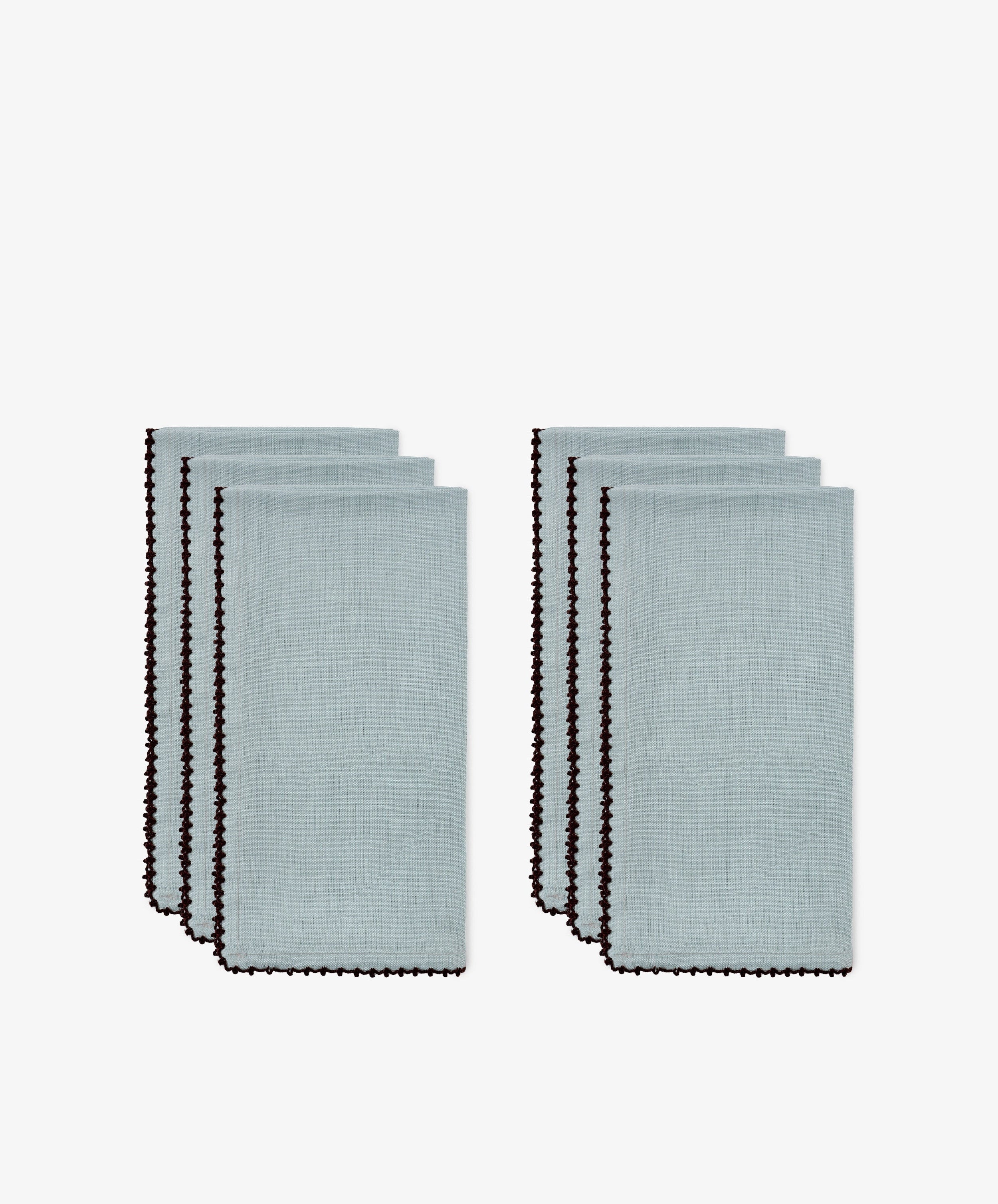 The Nico Napkin, Set of 6 in light blue linen are neatly folded and arranged in two separate stacks, each featuring a bold contrast stitch along the border.