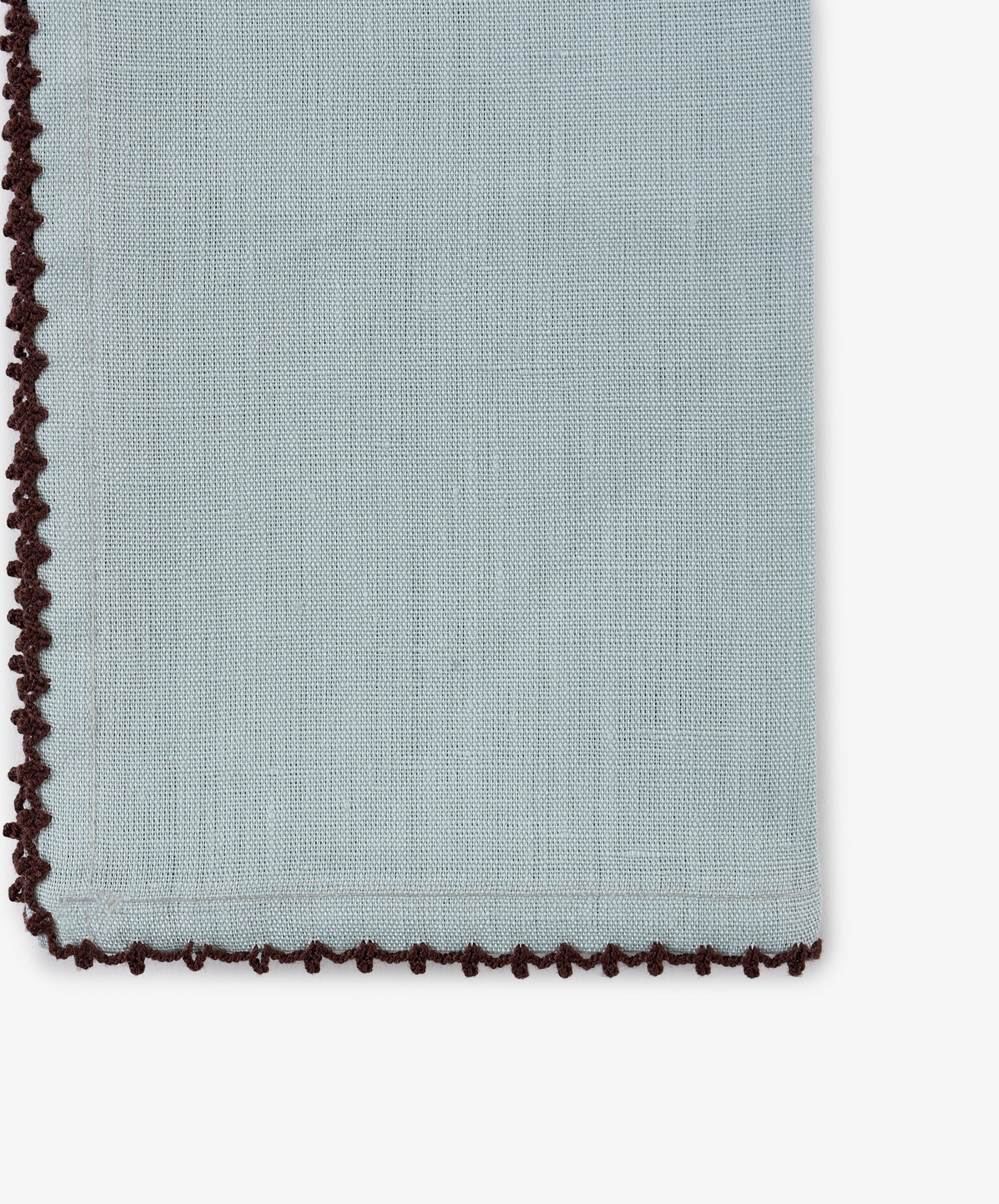 The Nico Napkin, Set of 6, is crafted from light blue linen and accented with a dark brown decorative crochet edge and a subtle contrast stitch. Shown here in close-up, the focus highlights the details at the bottom left corner.