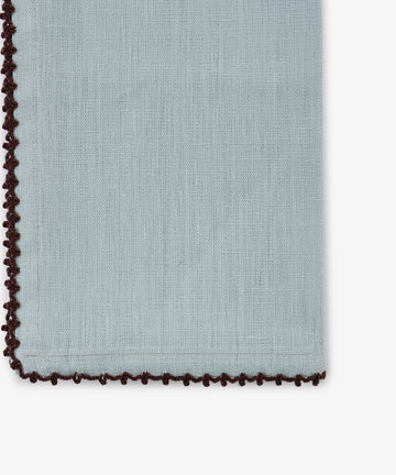 The Nico Napkin, Set of 6, is crafted from light blue linen and accented with a dark brown decorative crochet edge and a subtle contrast stitch. Shown here in close-up, the focus highlights the details at the bottom left corner.
