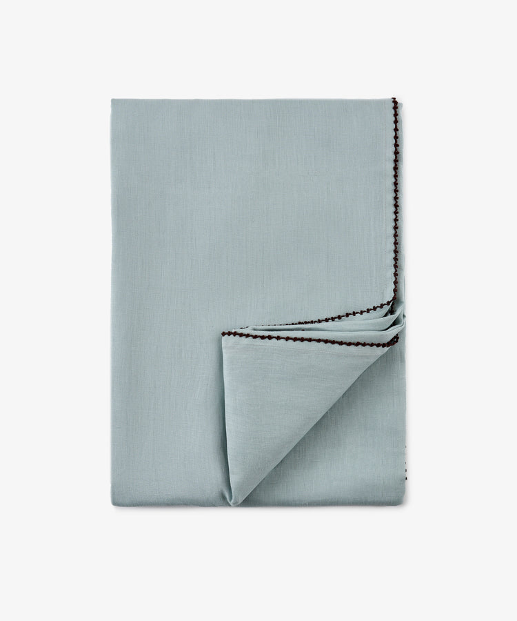 A neatly folded light blue Nico Tablecloth with dark stitched edging, reminiscent of a hand-finished table linen, shown on a plain white background.