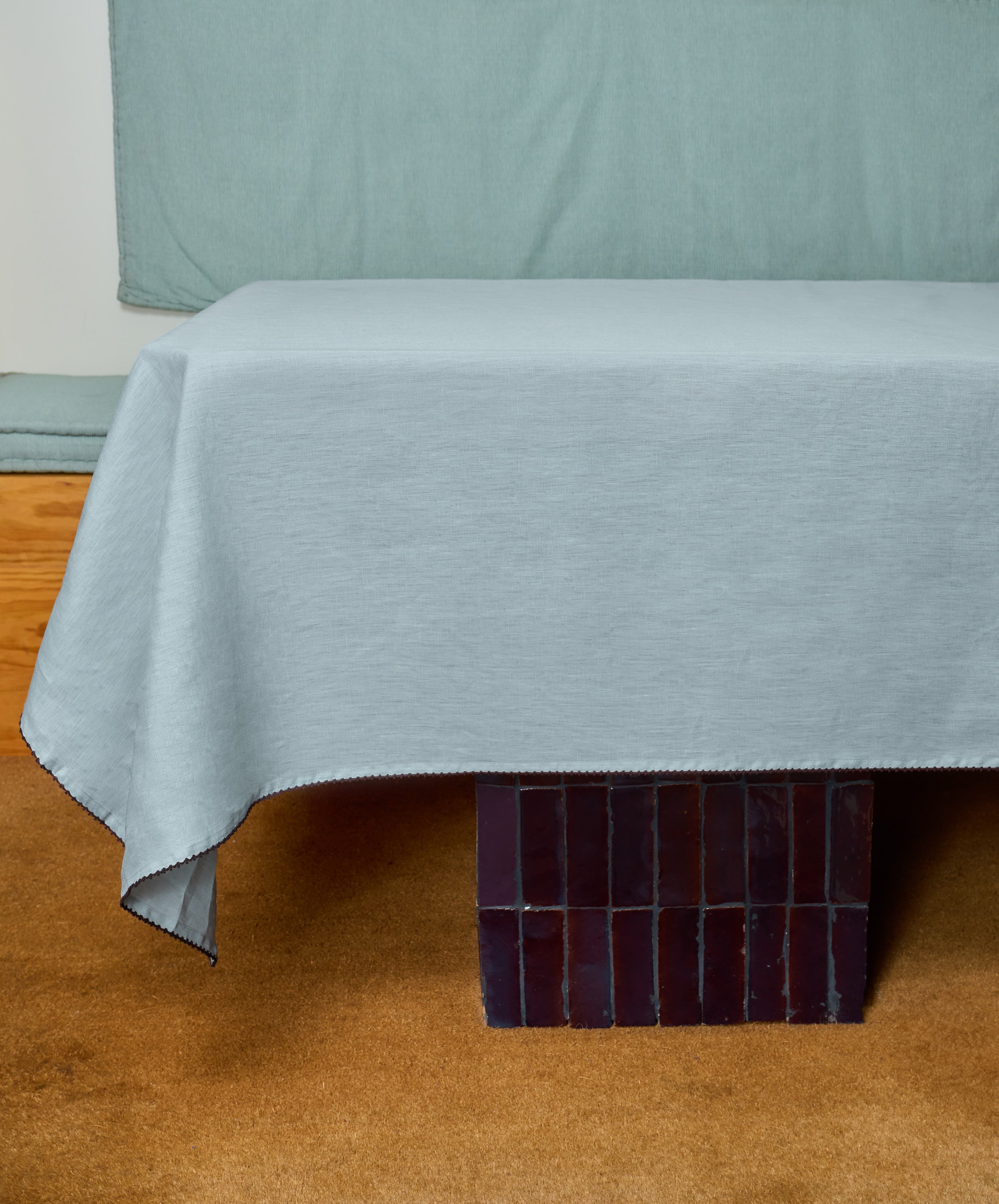 A table draped with the Nico Tablecloth in light gray linen rests on a carpeted floor, with dark rectangular tiles visible beneath its legs.
