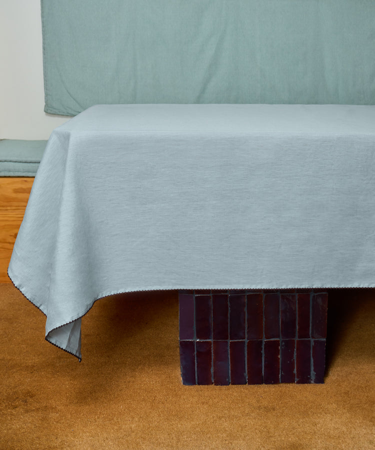 A table draped with the Nico Tablecloth in light gray linen rests on a carpeted floor, with dark rectangular tiles visible beneath its legs.