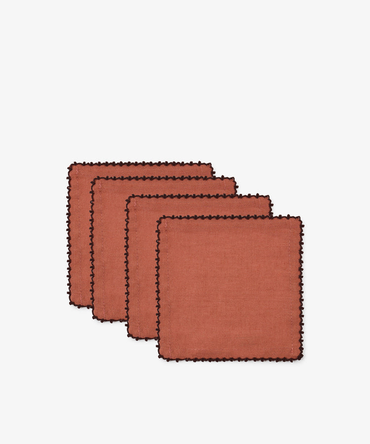 Set against a plain white background, the Nico Cocktail Napkin, Set of 4 features rust-colored linen napkins with black scalloped trim and subtle espresso contrast stitching, arranged in a staggered stack. Each hand-finished napkin brings an elegant touch to any table setting.