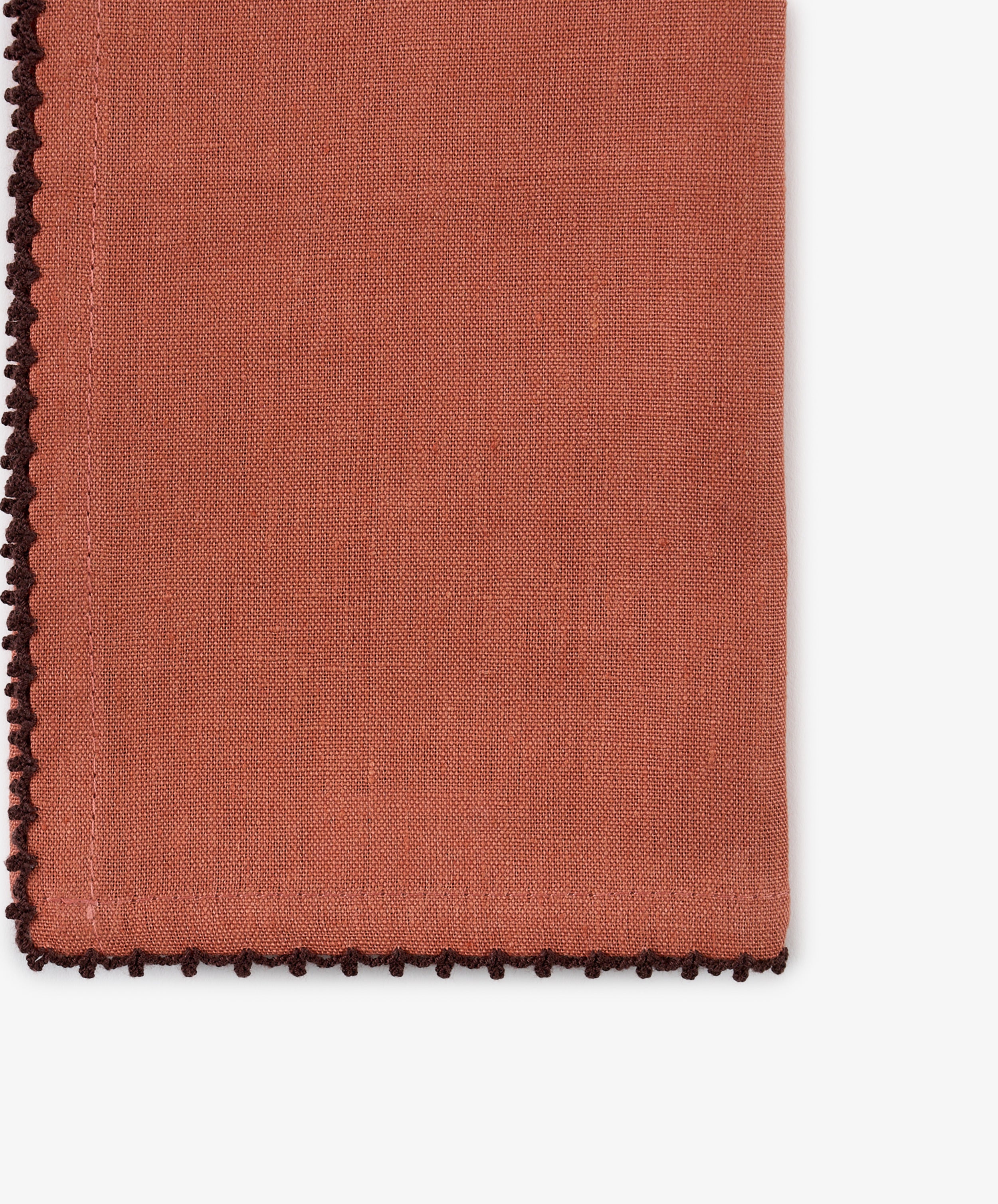 A close-up of a folded Nico Napkin from the set of 6 in rust linen, featuring decorative dark brown contrast stitch edging, beautifully displayed on a white background. This hand-finished napkin adds an elegant touch to any table setting.
