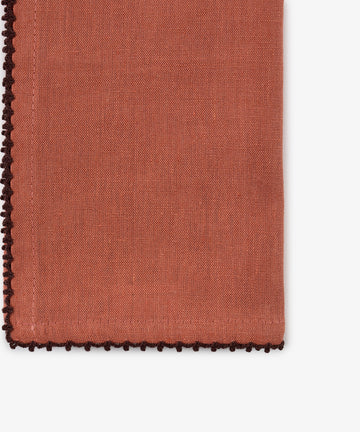 A close-up of a folded Nico Napkin from the set of 6 in rust linen, featuring decorative dark brown contrast stitch edging, beautifully displayed on a white background. This hand-finished napkin adds an elegant touch to any table setting.