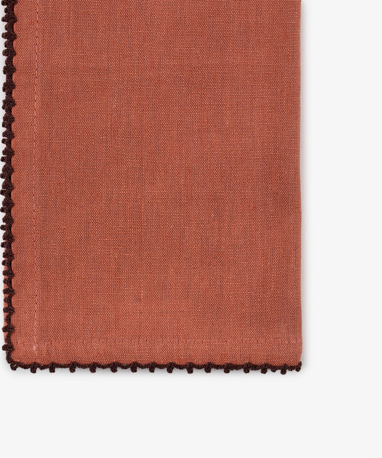 A close-up of a folded Nico Napkin from the set of 6 in rust linen, featuring decorative dark brown contrast stitch edging, beautifully displayed on a white background. This hand-finished napkin adds an elegant touch to any table setting.