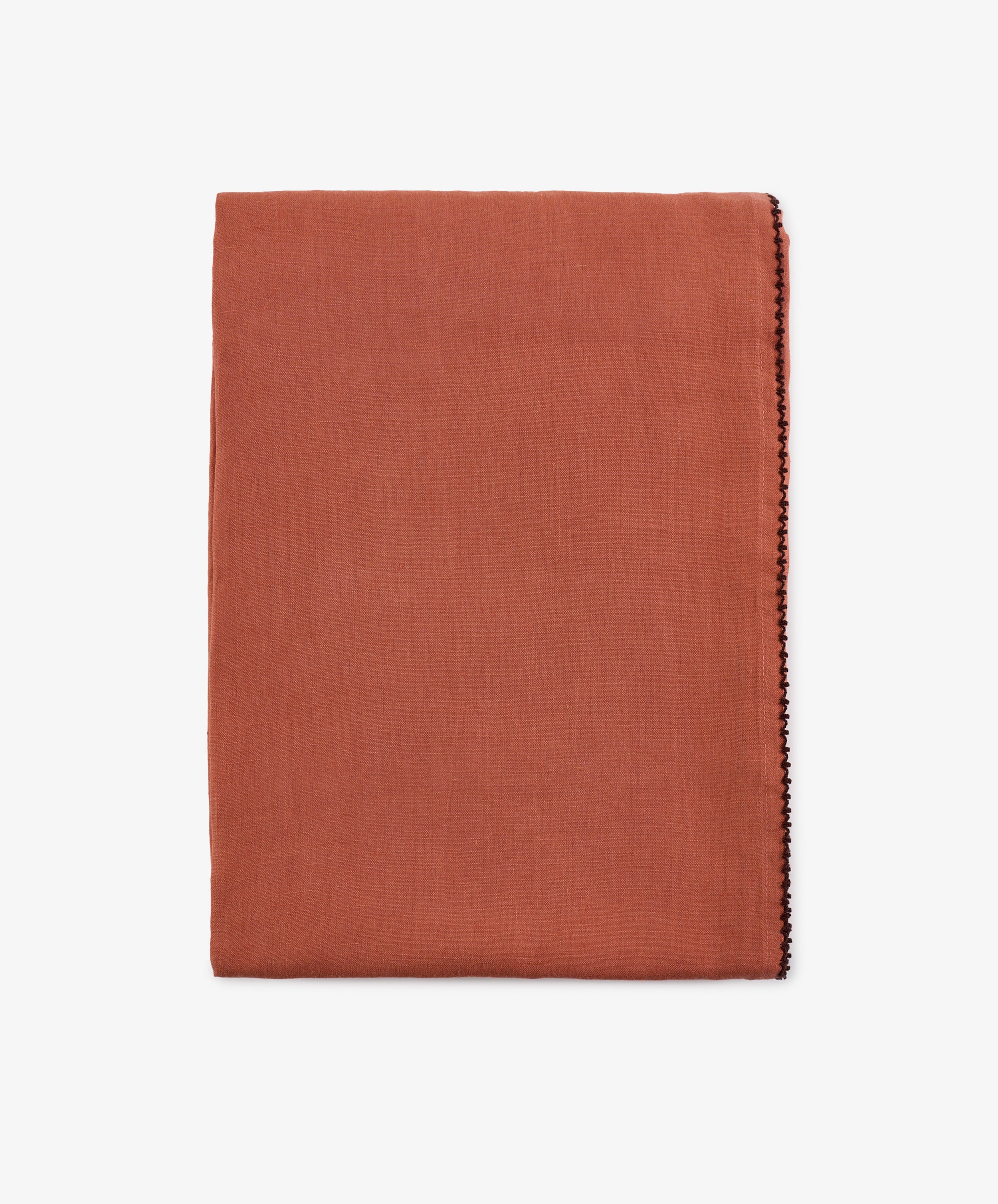 A folded rust linen Nico Tablecloth with a thin decorative stitch along one edge, set against a plain white background. This hand-finished detail enhances the tablecloth’s elegant simplicity.